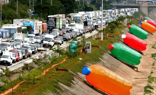 obra de eduardo srur. mostra garrafas de plástico gigantes acostadas ao lado de uma rodovia movimentada, ilustra a arte com reciclagem